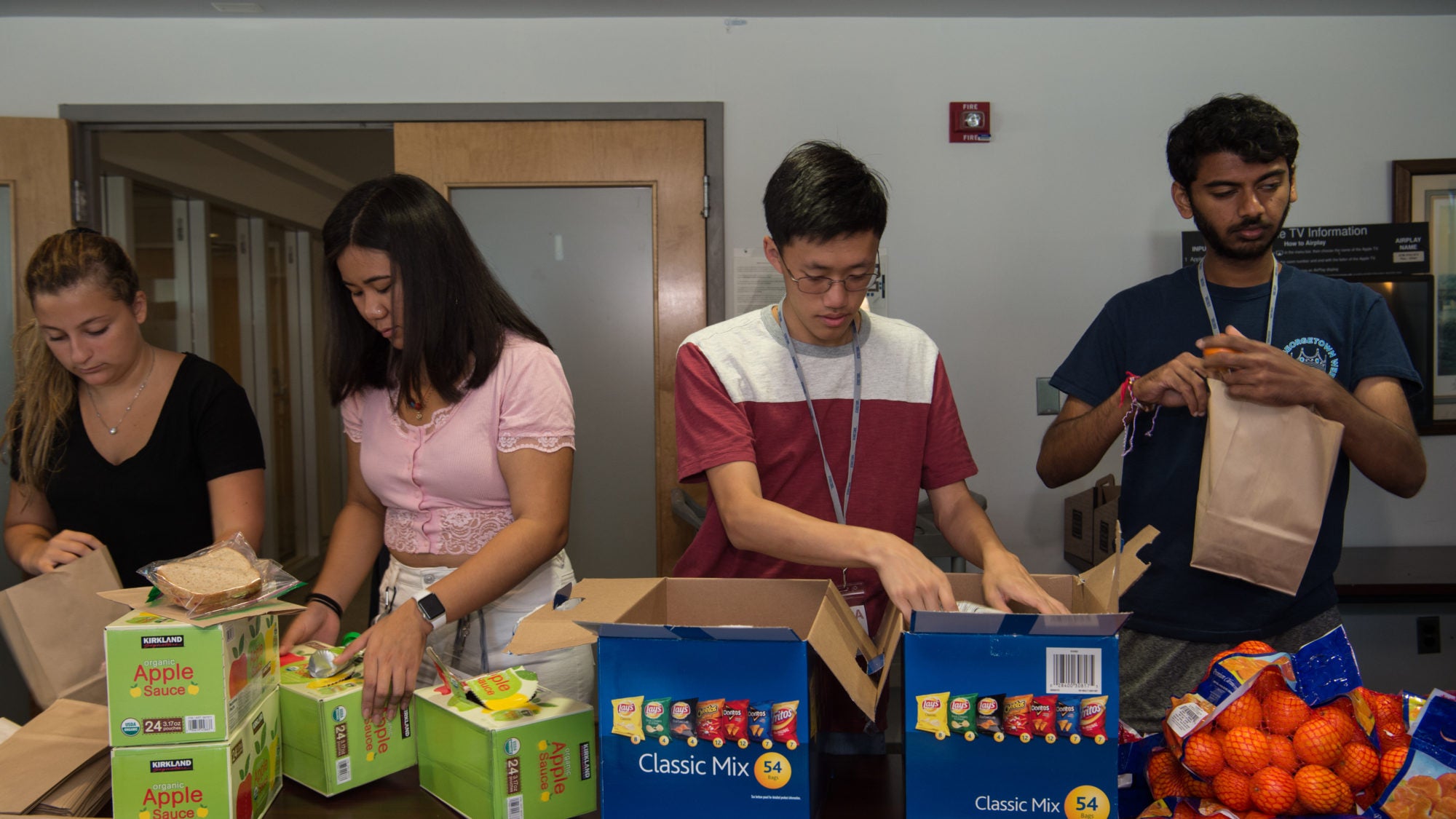 Photo shows four students packing lunch bags in an assembly line.