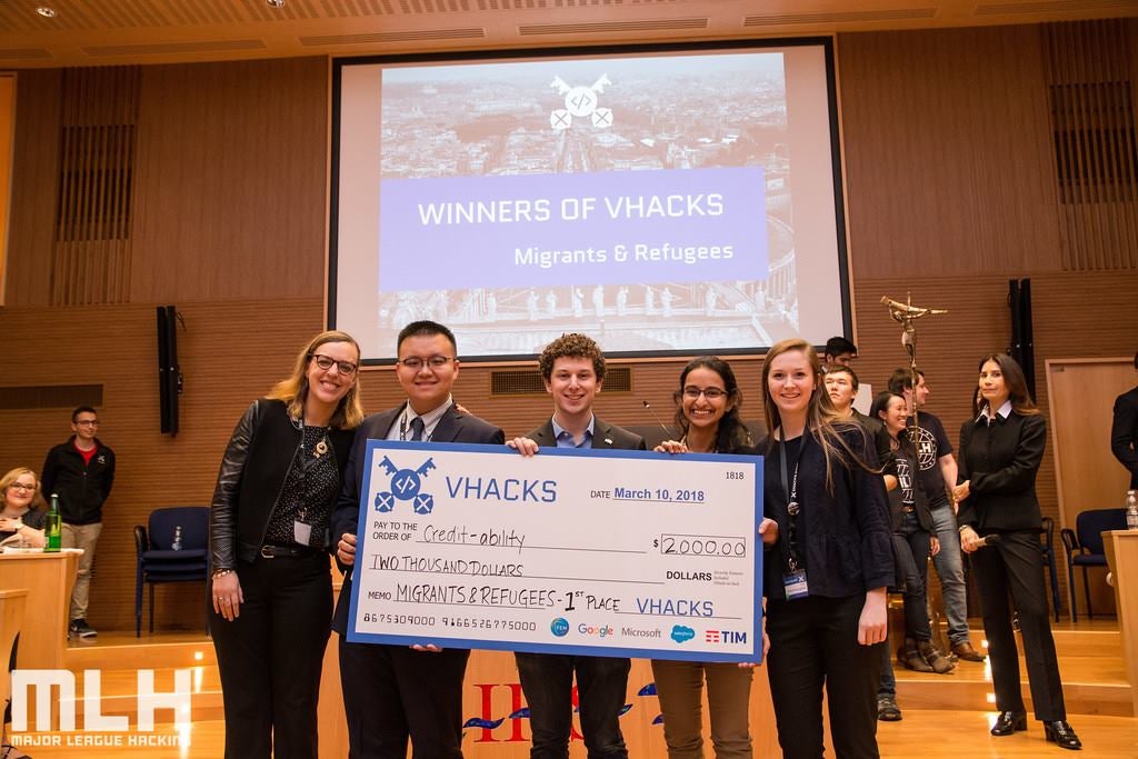 Roisin McLoughlin, Jake Glass, Lucy Obus, Rushika Shekhar and Yanchen Wang stand on a stage holding an oversized check address to Credit-ability for $2,000 with other people and a screen behind them reading Winners of Hacks, Migrants and Refugees