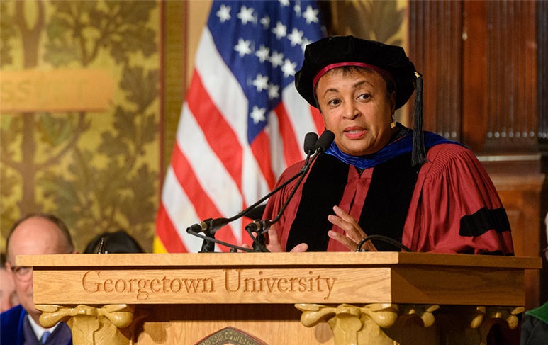 Carla Hayden at Georgetown University podium wearing cap and gown with American flag behind her