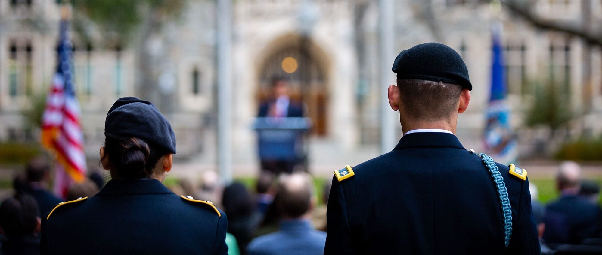 A female and male member of the military stand in uniform facing White-Gravenor Hall with an American flag in the foreground.