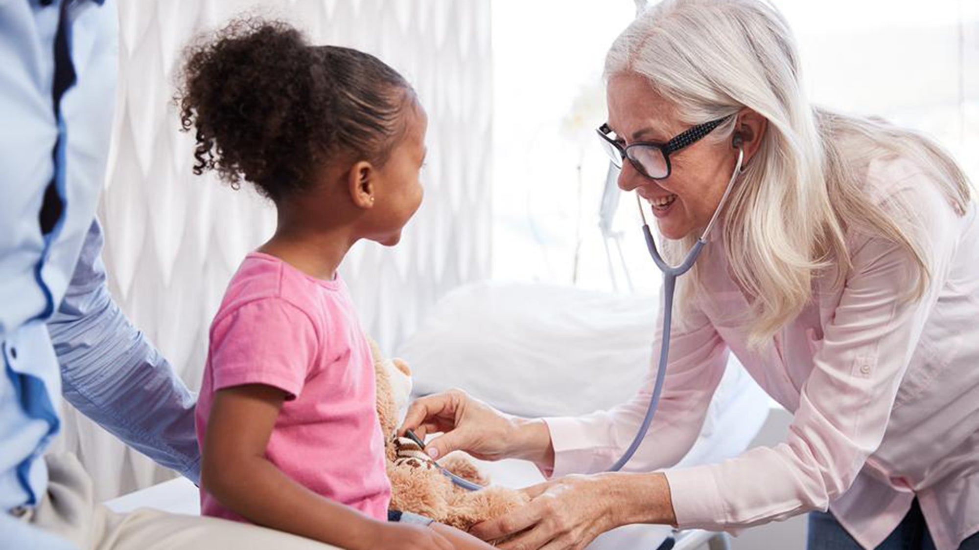 A little girl sits as her guardian stands behind her as a doctor listens to her pulse with a stethoscope.