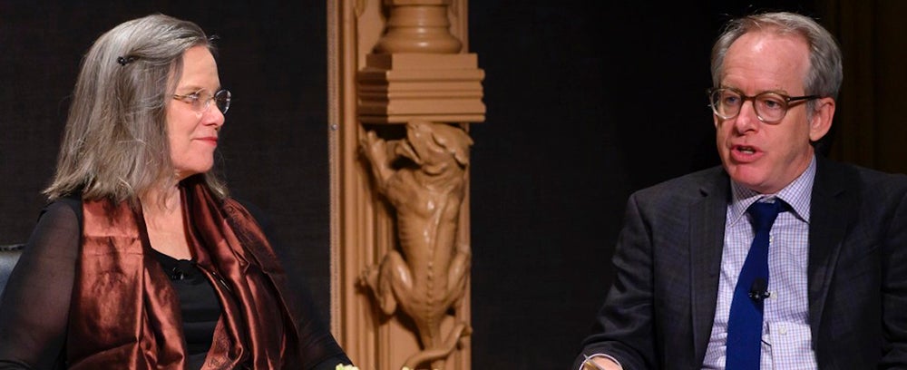 Carolyn Forché sits talking with Paul Elie with a wooden column between them with a wooden gargoyle on it