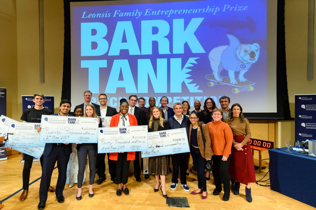 Students stand with oversized checks, Ted Leonsis and professors on a stage with a large screen reading Bark Tank and an illustration of Jack the Bulldog