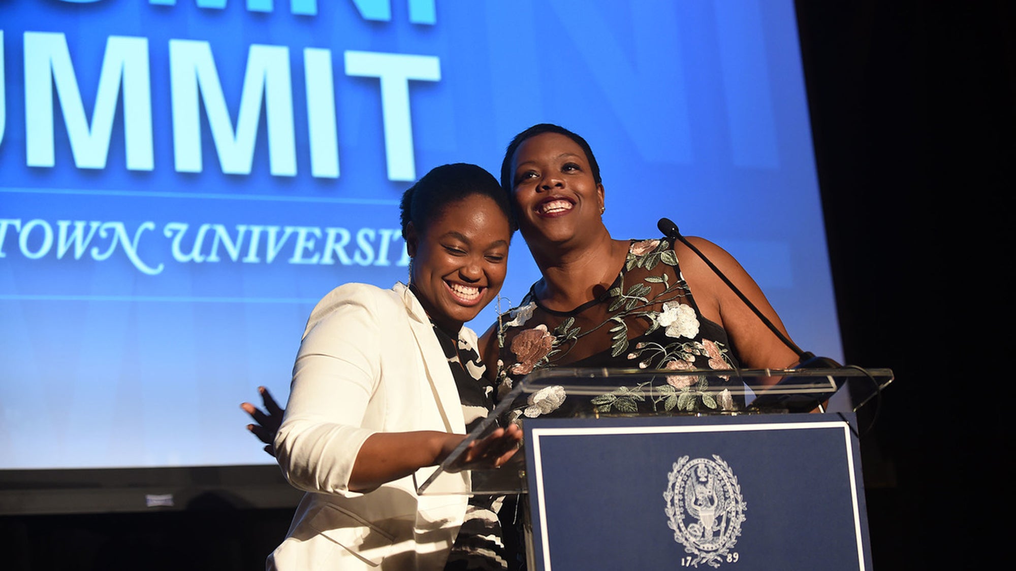 Elizabeth Nalunga and Kaya Henderson embrace at the lectern on stage with Black Alumni Summit signage on a blue background.