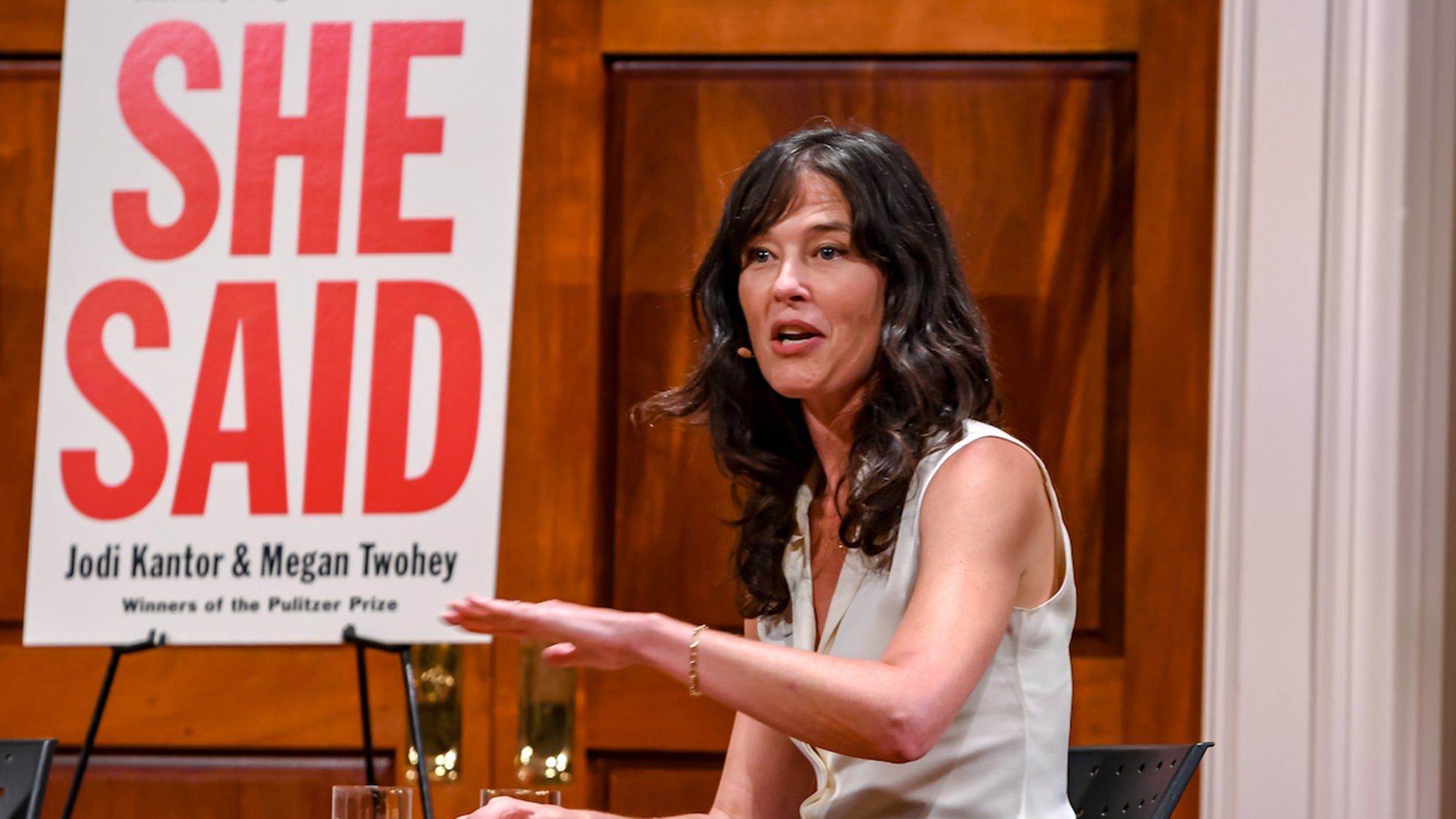 Megan Twohey gestures sitting onstage with a glass of water and a stand behind her with a poster that reads "She said, Jodi Kantor & Megan Twohey, Winners of the Pulitzer Prize. Photo by Ari Strauss