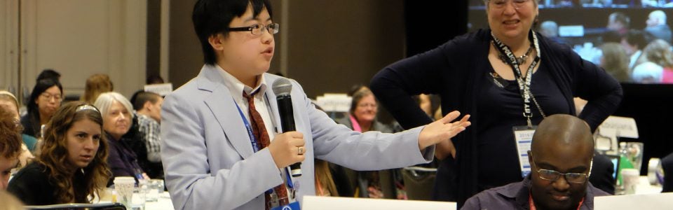 Lydia X.Z. Brown standing with a microphone with people sitting and standing around them.