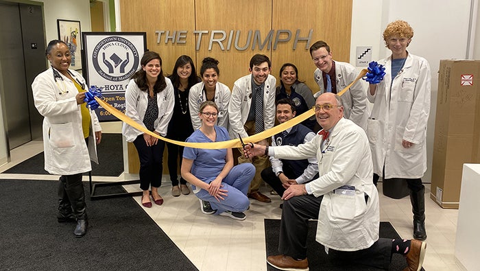 DDr. Stephen Ray Mitchell kneels on the floor cutting a yellow ribbon held by two other doctors as doctors and other people kneel or squat in front of a sign reading The Triumph