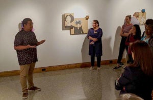 Kenlontaé Turner speaks to a group of visitors at a gallery with his artwork hanging in the background.