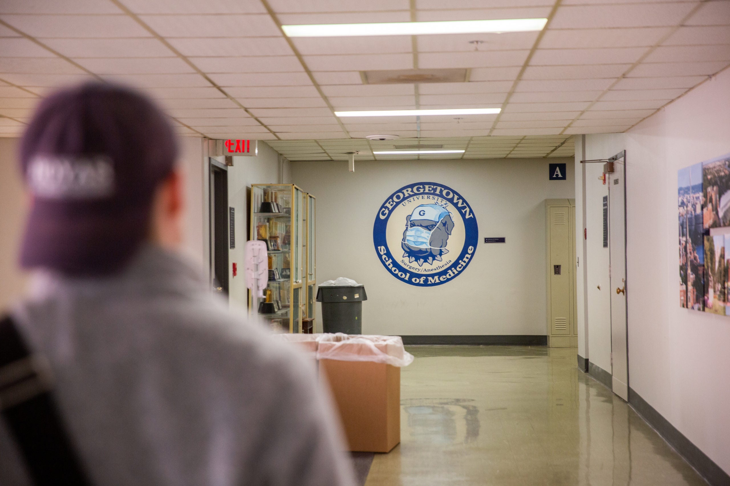 A student with a blue hat on backwards walks down a hallway, at the end of which a logo for the School of Medicine is painted on the wall.