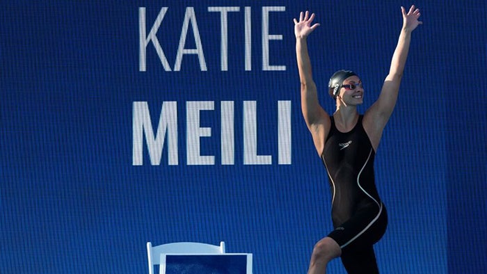 Katie Meili in swimsuit and bathing cap with her arms raised in front of a banner with her name on it