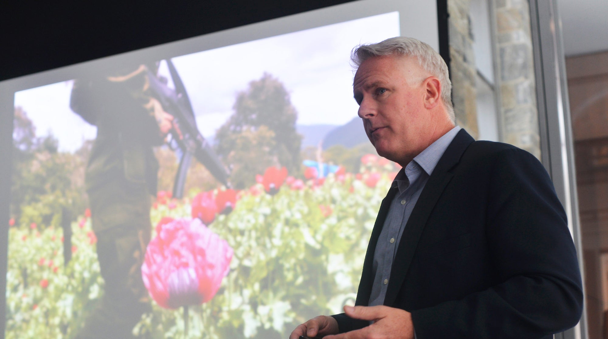 John Moore standing in front of a screen with his one of his photo, this one of a man standing in a field with flowers and holding a semi-automatic weapon