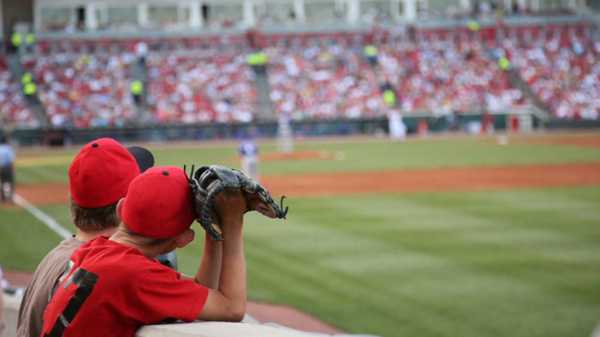 Two boys wearing baseball hats and gloves turn their heads toward the baseball field to watch a game being played.