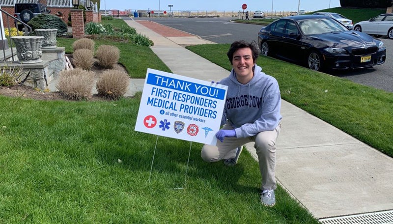 Liam Marshall squatting on a front lawn with a sign stuck in the lawn reading "Thank You First Responders and Medical Providers and all other essential workers."