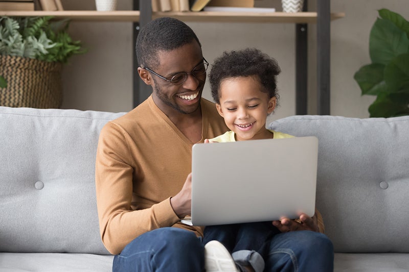 A father sits with a daughter on a sofa with the daughter holding a laptop, both are smiling