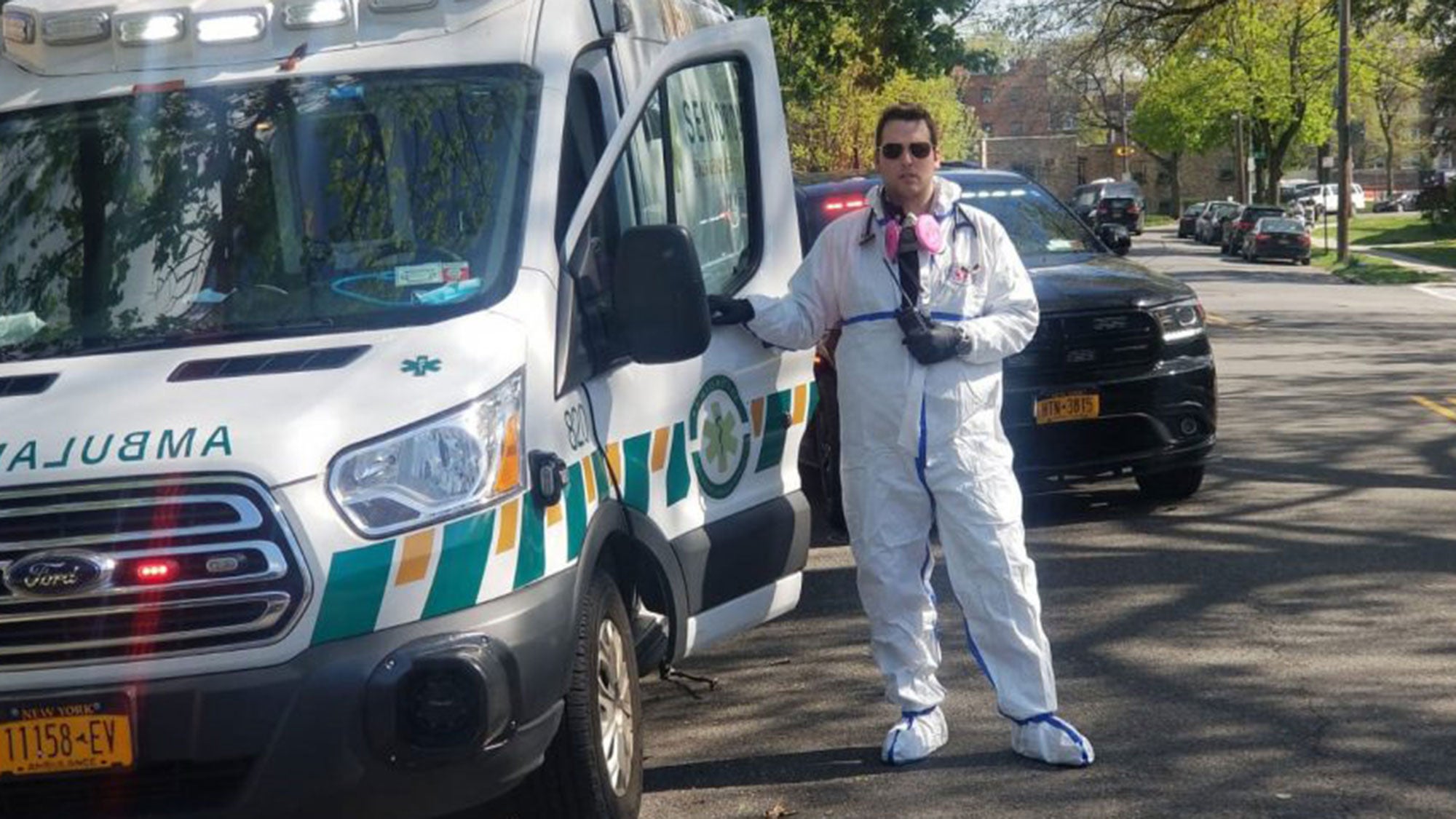 Mark Vatch stands near an emergency medical vehicle dresses in a protective white suit and mask around his neck.