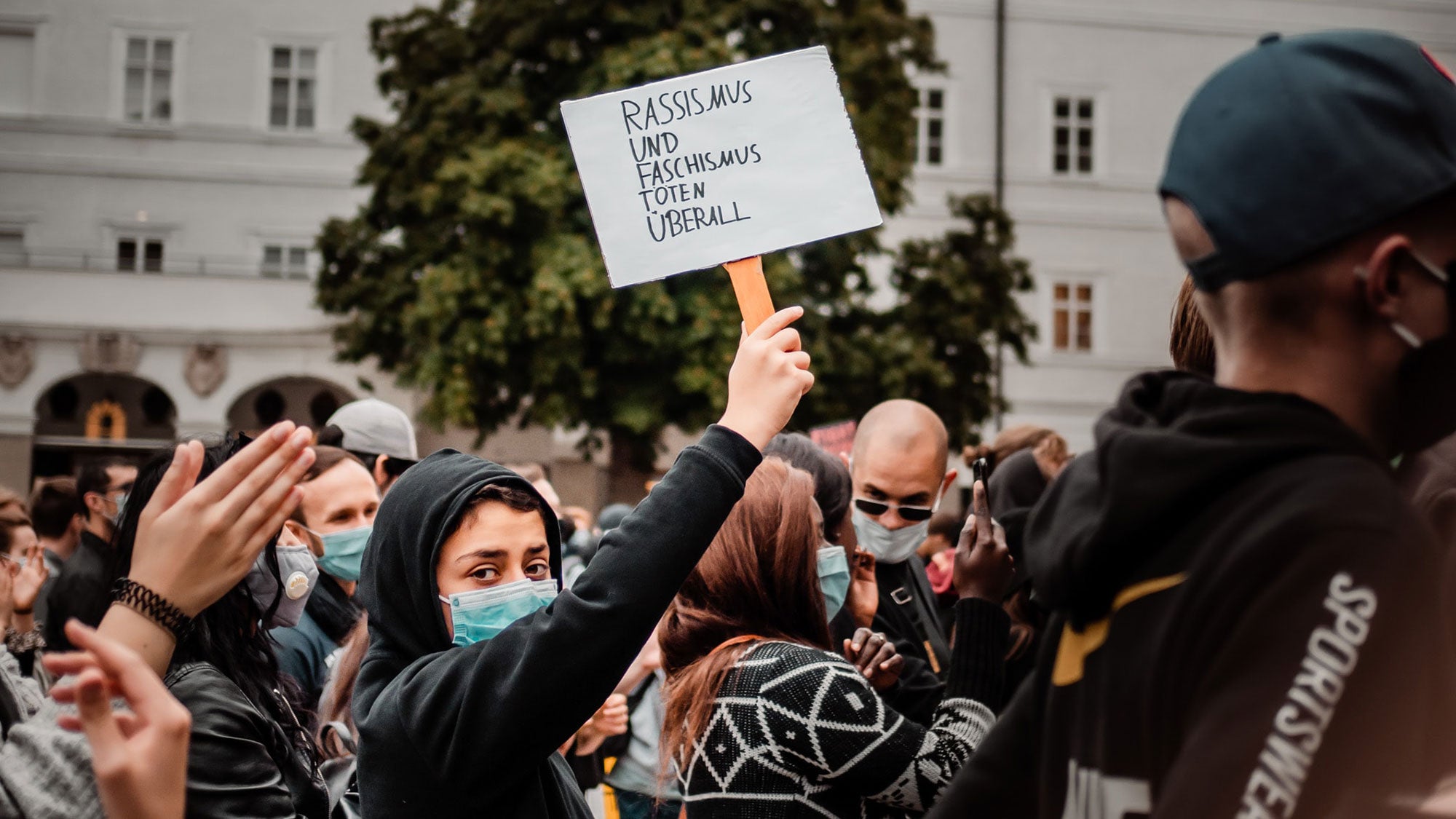 A protester stands in a crowd wearing a face mask and holding a sign that says "Racism and facism kill everywhere."