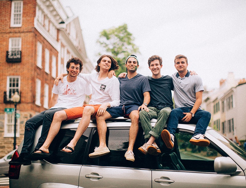 Five young men sit on top of a car with buildings behind them