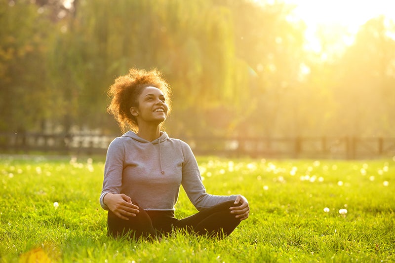 A woman sits in a field crossed-legged with the sun shining on her smiling face and trees in the background