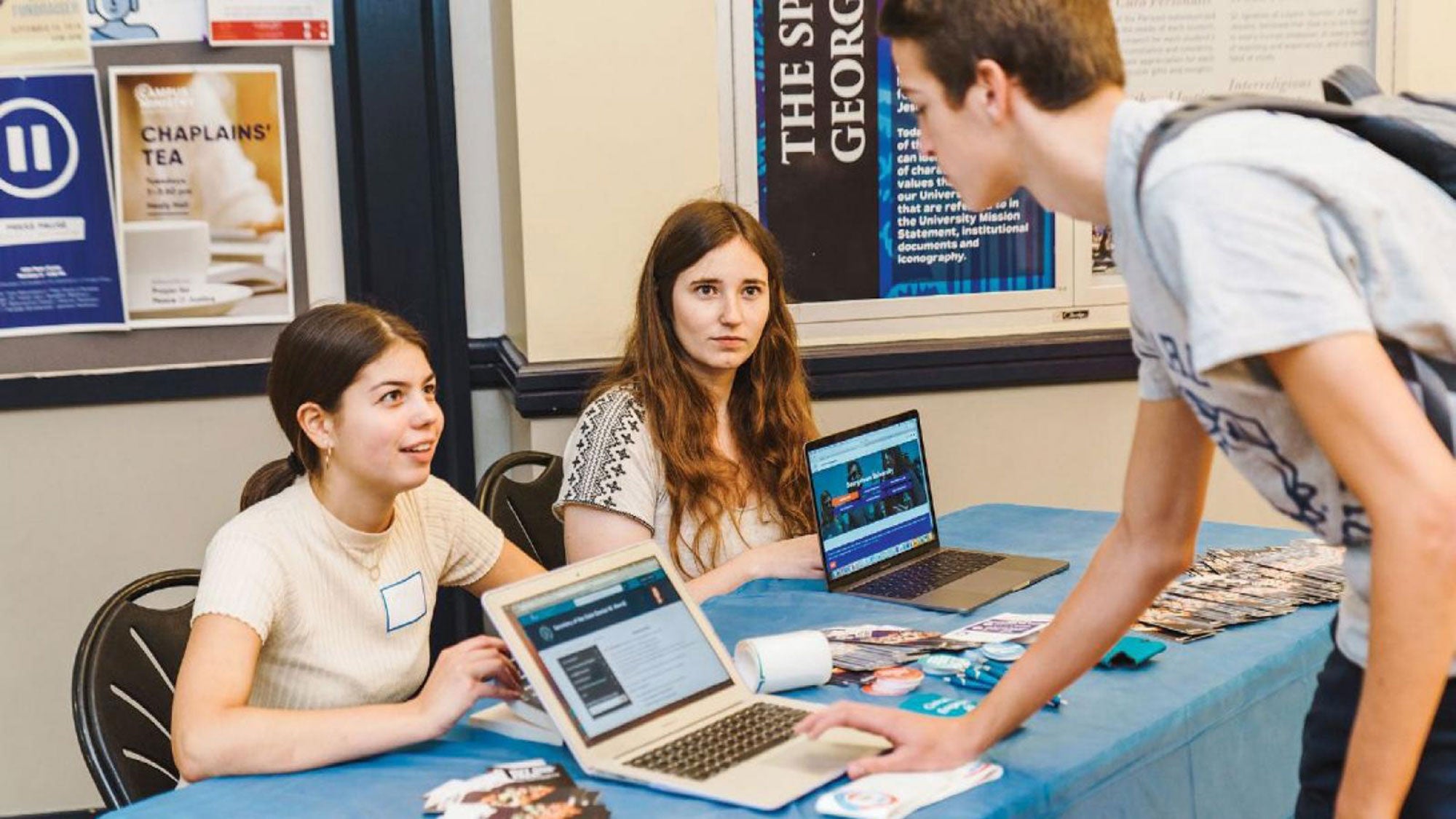 Two students sit at a table while another student registers to vote on a laptop at the table.