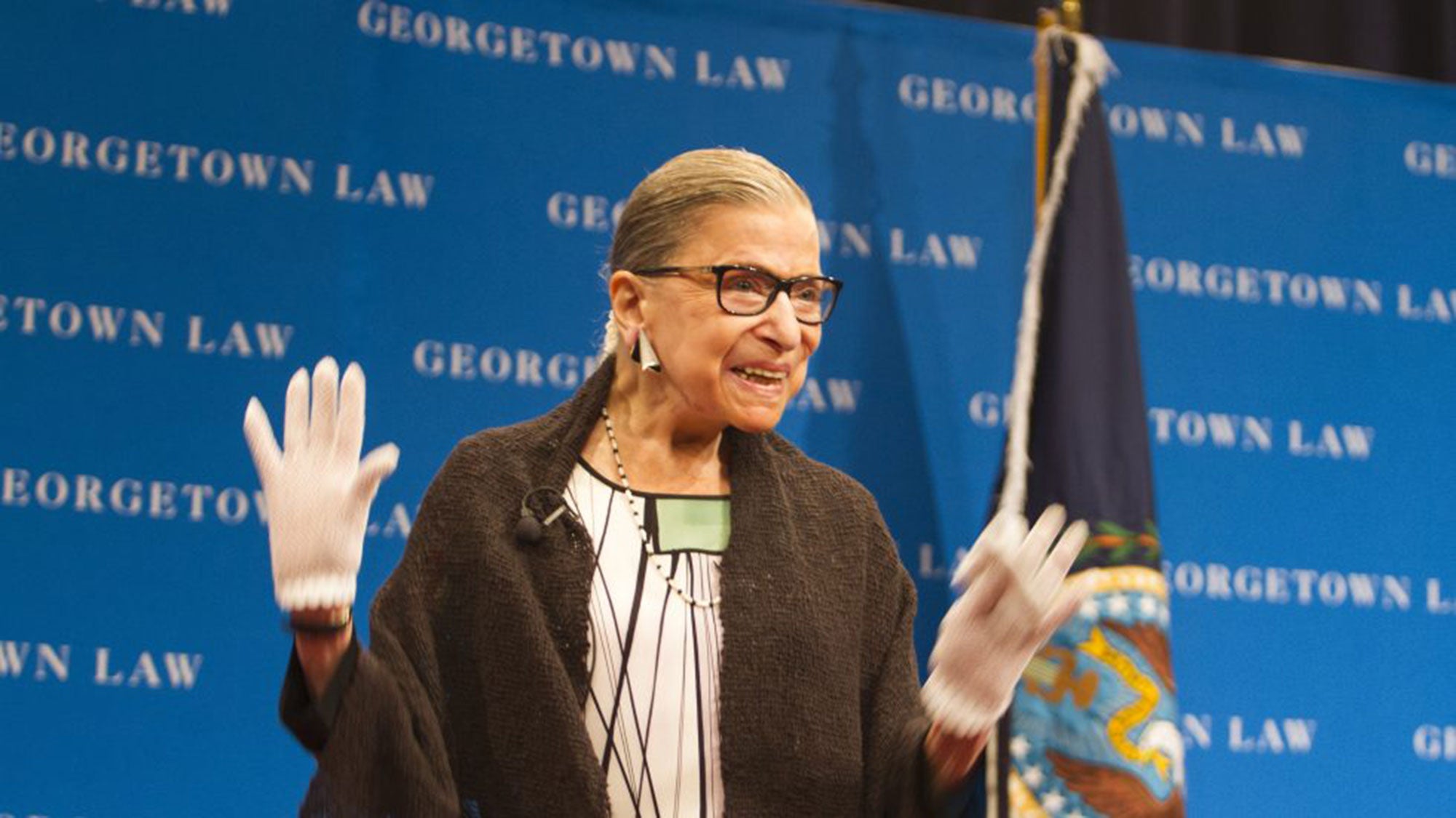 Ruth Bader Ginsburg stands on stage waving to the audience with both hands with a Georgetown Law background.