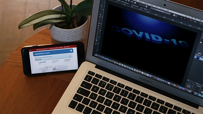 A computer open on a desk with the words COVID-19 and smartphone and plant next to it.