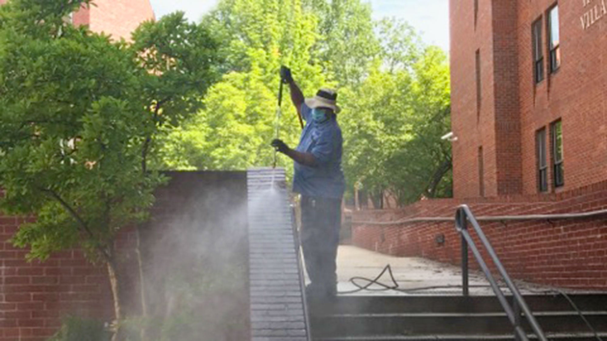 A man wears a mask while pressure washing the brick around stairs on campus.