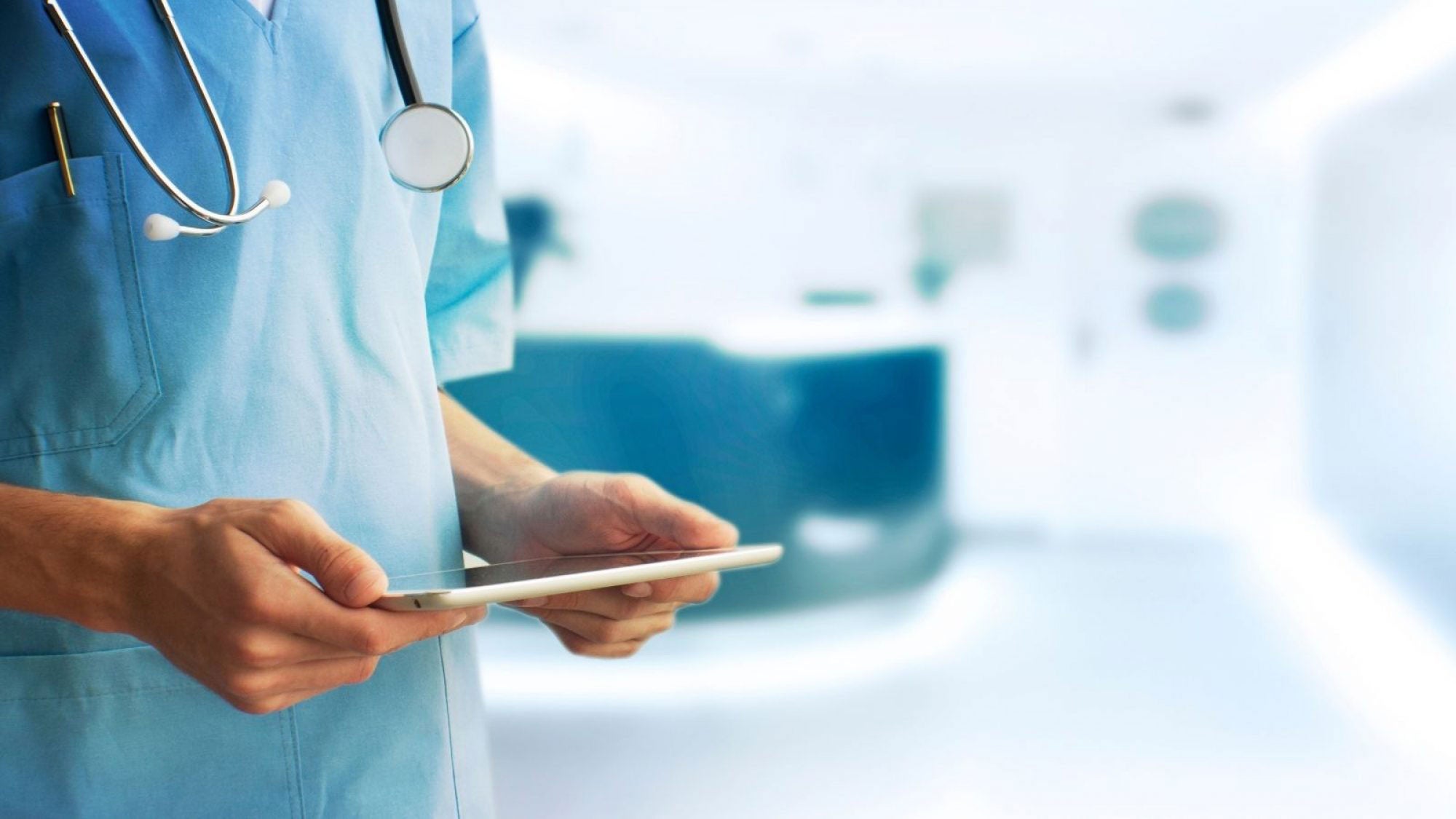 A healthcare worker in blue scrubs looks down at a hanheld device.