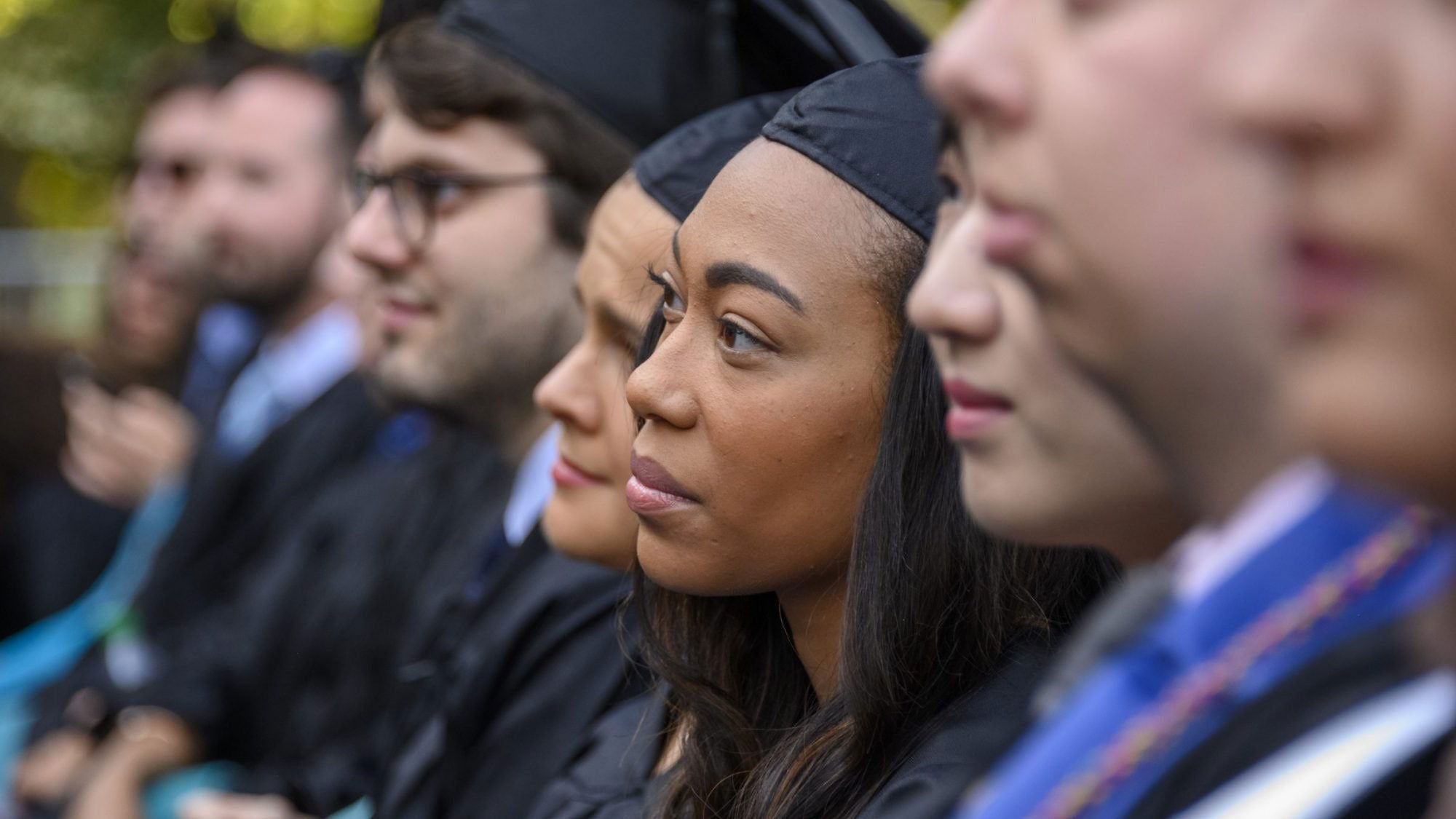 Students dressed on academic regalia sit and listen to someone speaking.