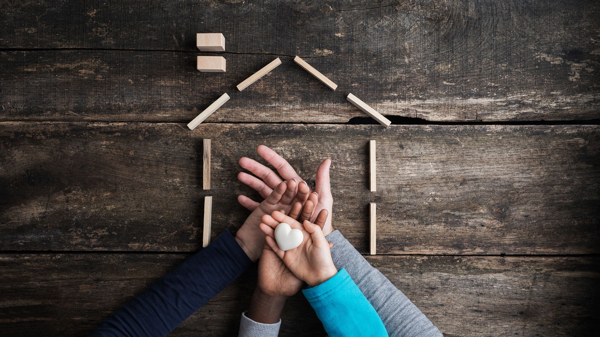 The hands of a family hold a heart-shaped item with hands placed in the center of a outlined house-shaped structure.