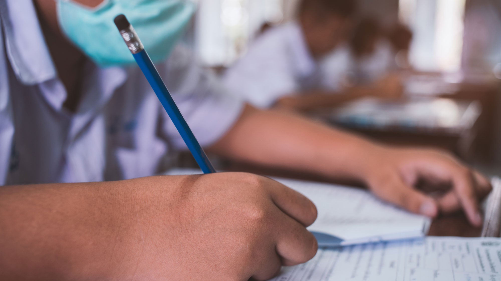 A student wears a mask as he writes on paper while sitting at a desk.