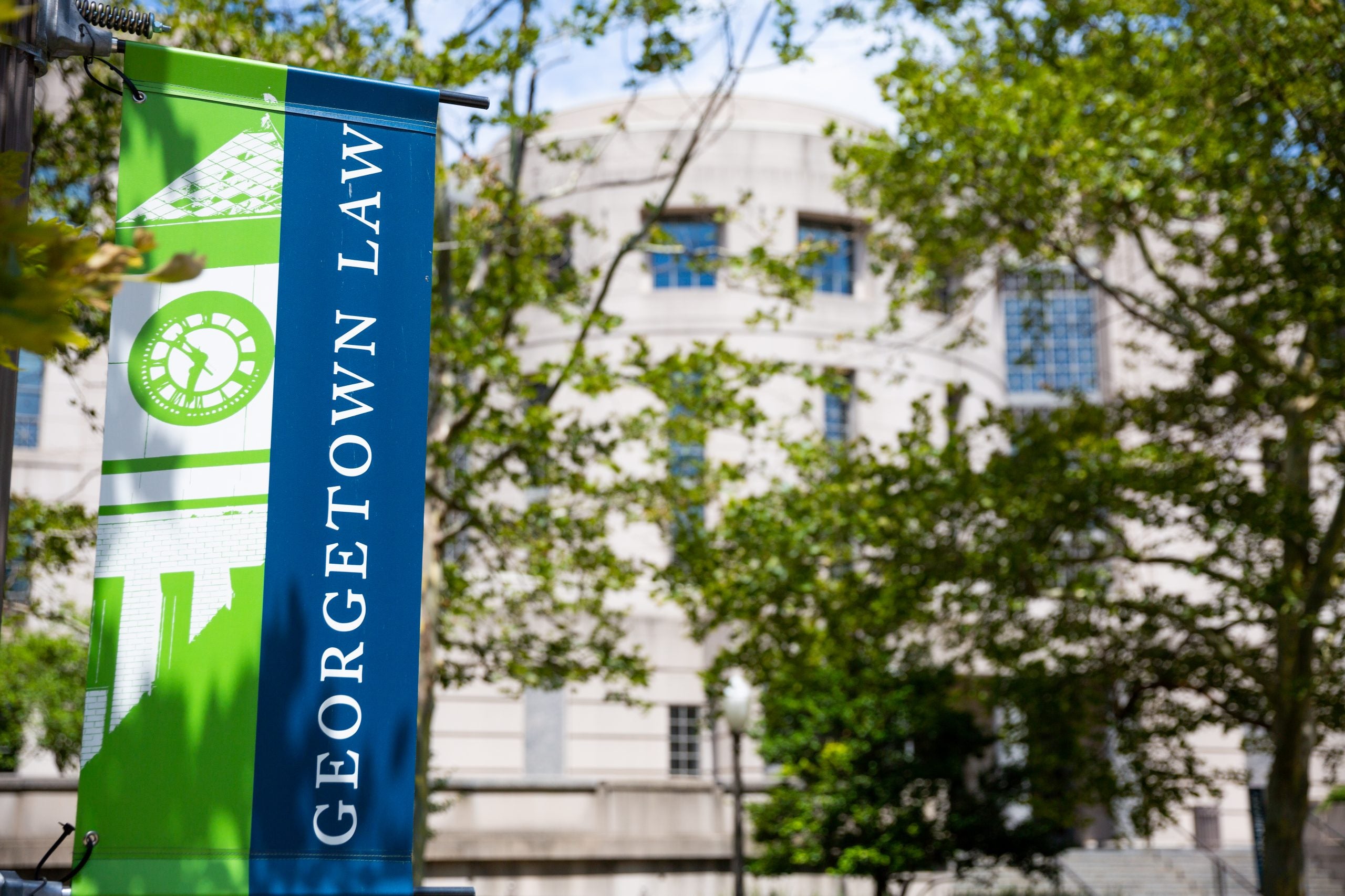 An image of a blue flag (left) with the words "Georgetown Law" in front of a white Georgetown Law building with green trees surrounding it.