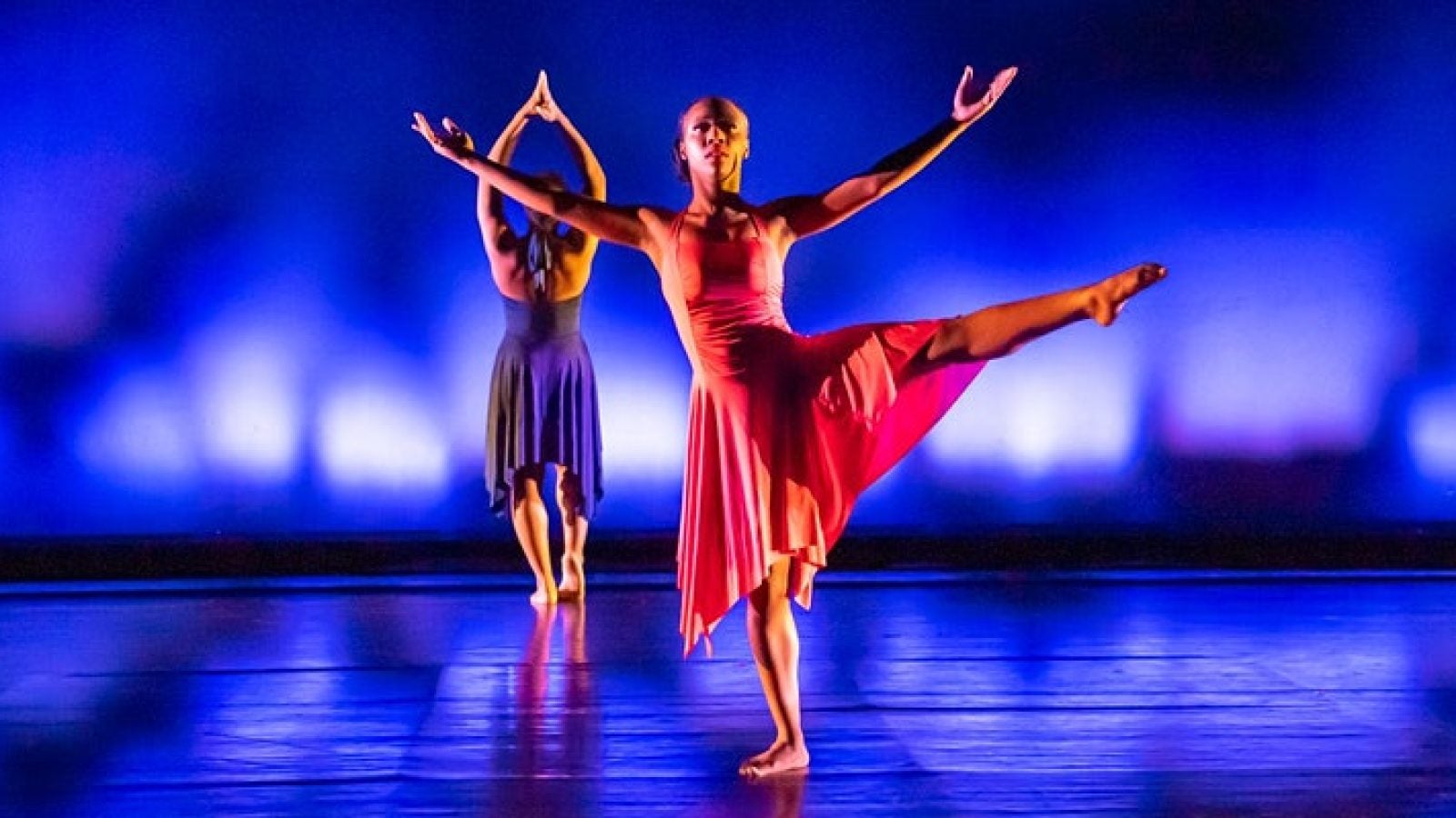 Two dancers from Black Movements Dance Theatre perform, one in a blue dress with hands clasped above her head, and one in a red dress, with her left leg and arms outsretched