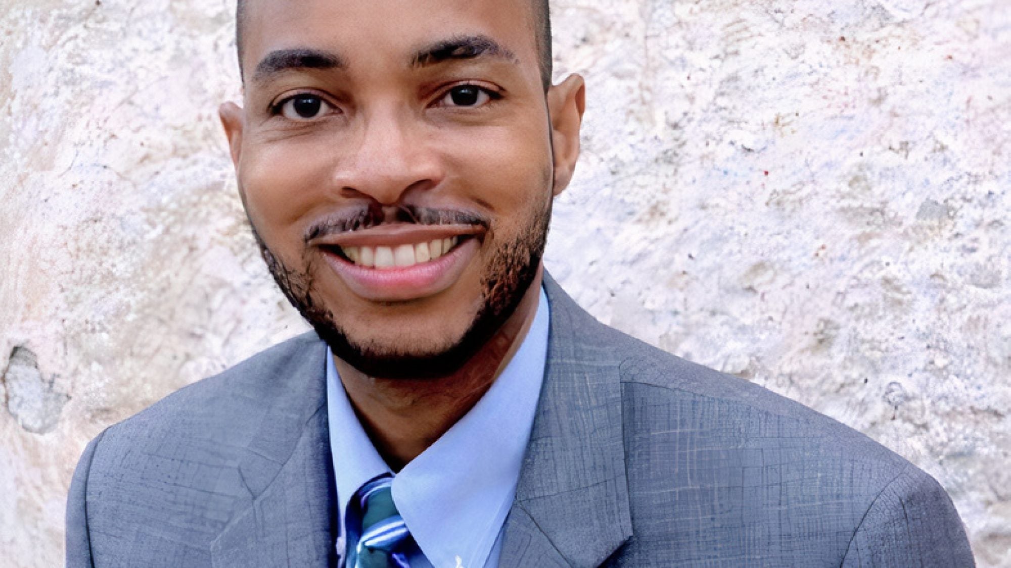 Melvin L. Butler headshot in gray suit with stone background.