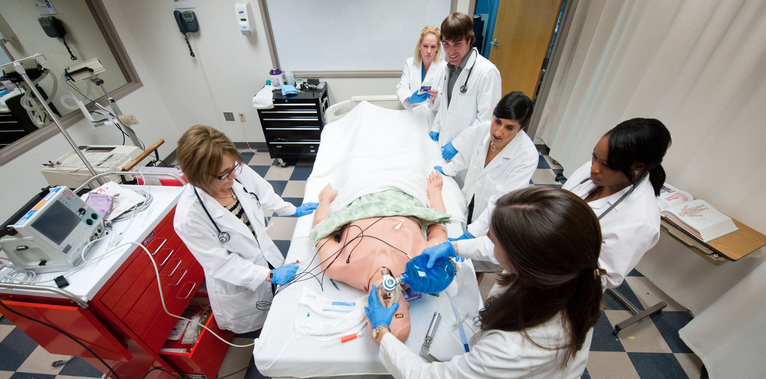 Students and professors in white coats around a fake body