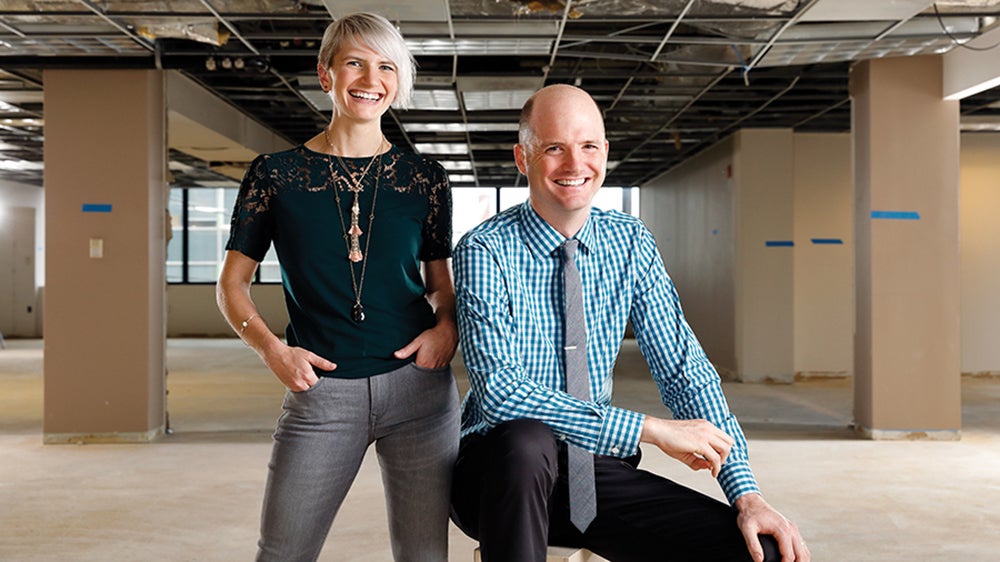 One alumni, dressed in a black shirt and jeans, stands next to another business school alumni, who's dressed in a blue shirt, a tie and black pants.