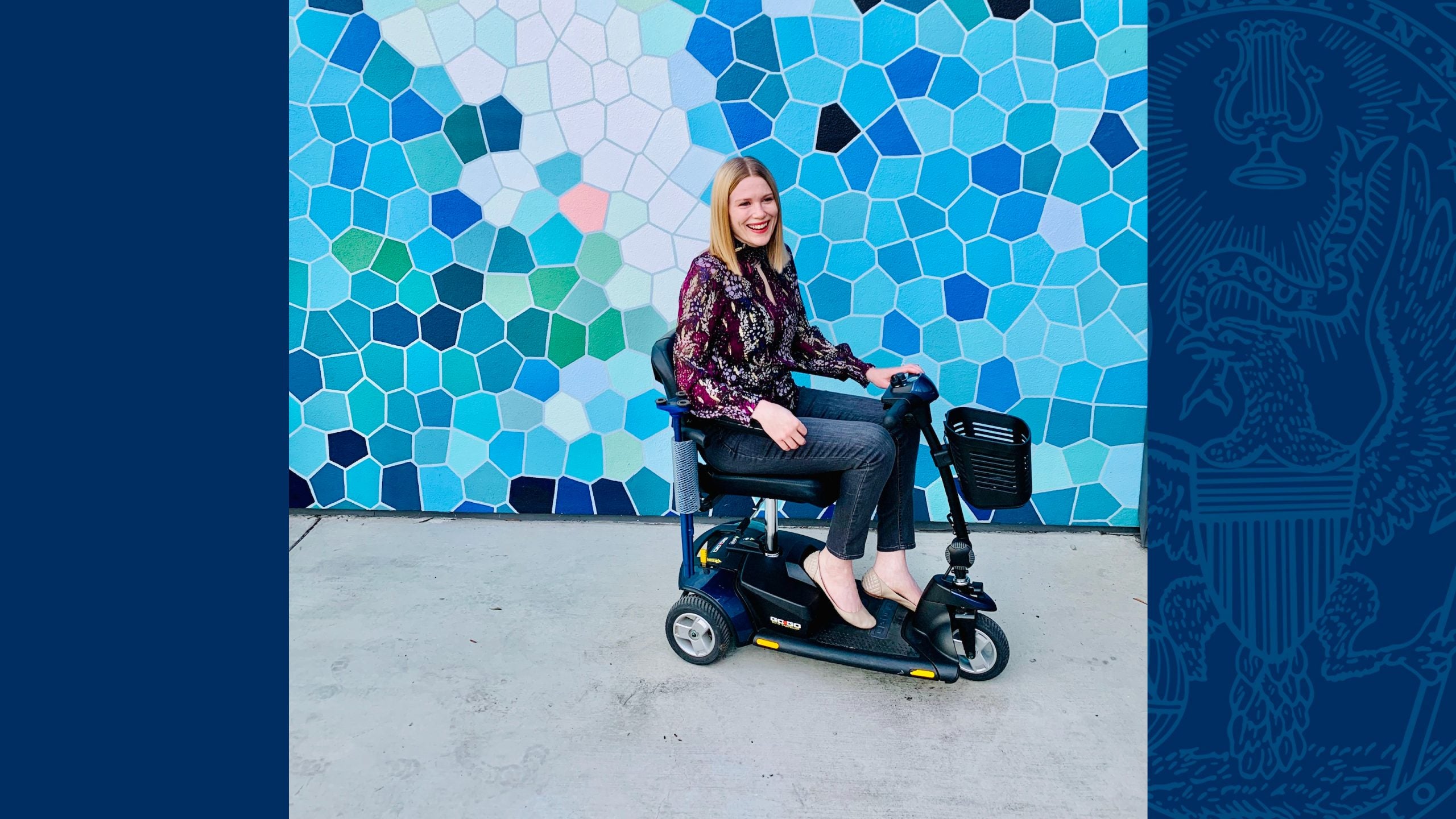 A white woman with straight, blonde hair, wearing a burgundy patterned shirt and grey jeans sits on a blue and black mobility scooter and smiles in front of a wall with a variety of blue and white geometric shapes