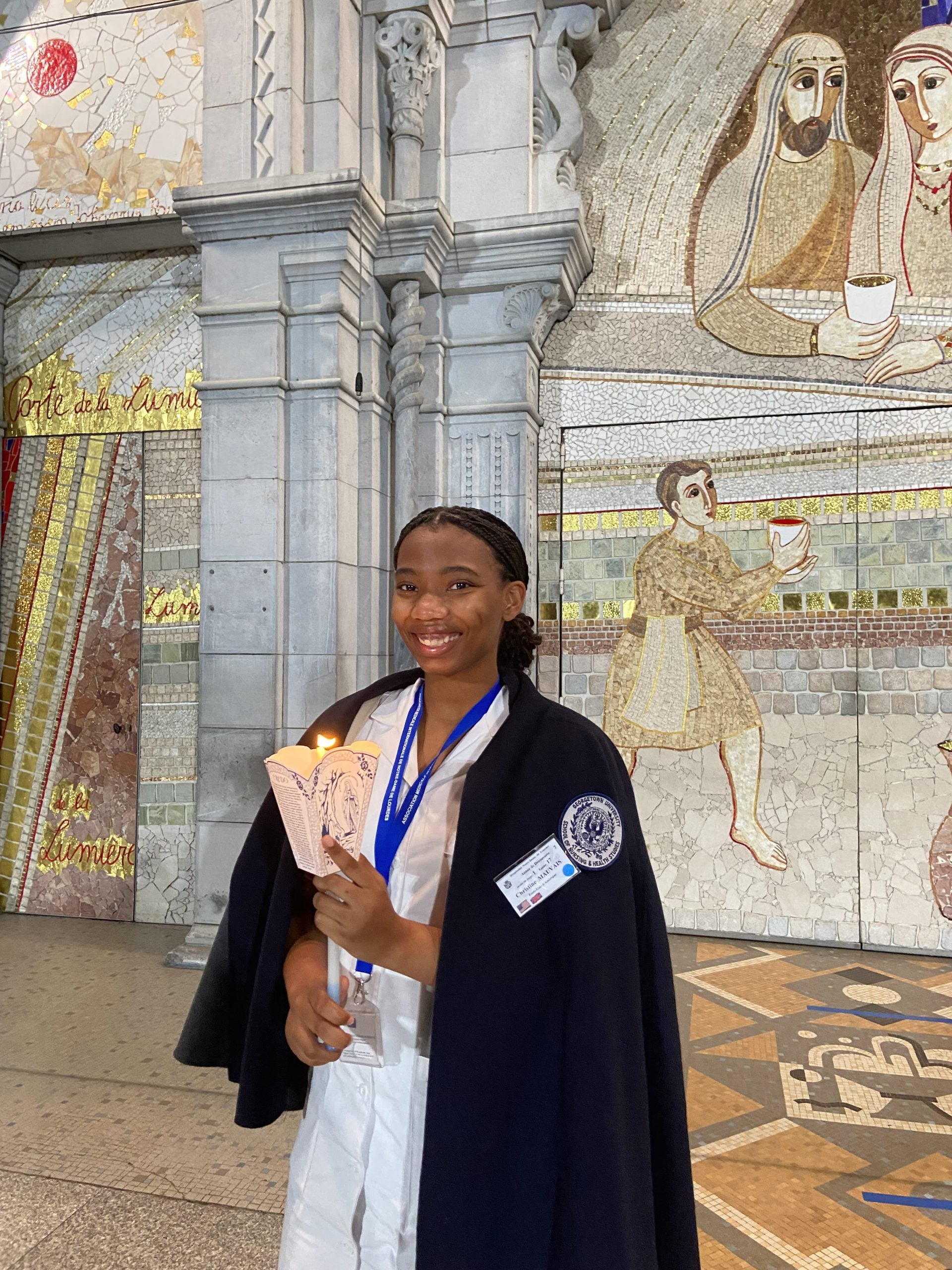 Christine Mauvais wears a white dress, blue medal and black coat off her shoulders while holding a candle in front of mosaic religious art