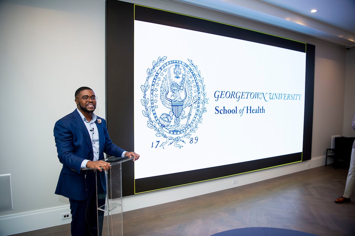 Christopher King at a clear podium in front of a projection with the text "Georgetown University School of Health"