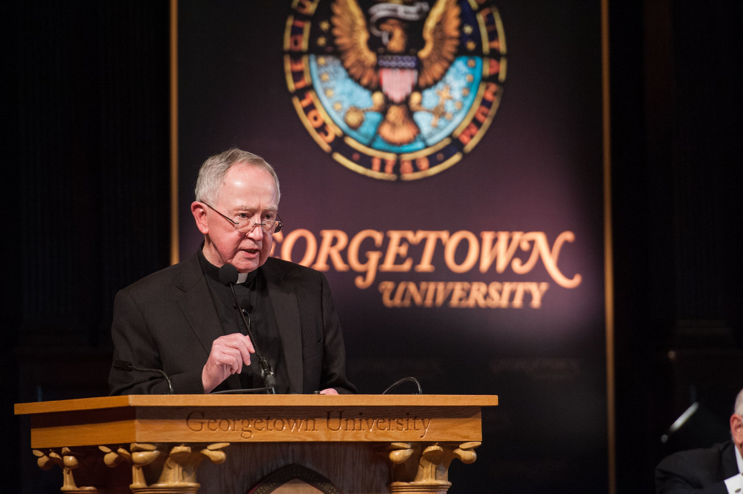 Fr. O'Malley wears glasses, a priest's collar and black clothes while giving a speech in front of a Georgetown University sign