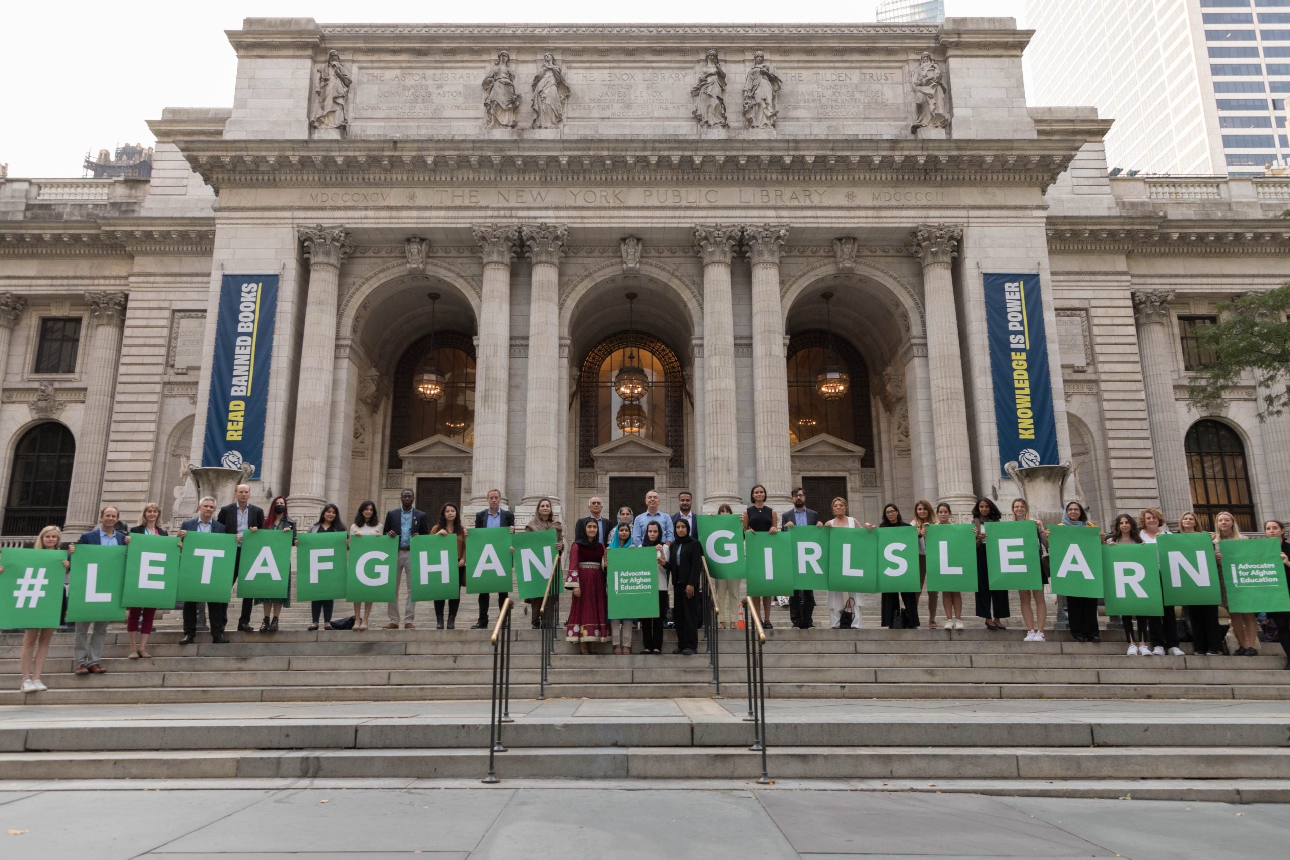 Women holding individual signs with green letters in front of an old building spelling out "Let Afghan Girls Learn"