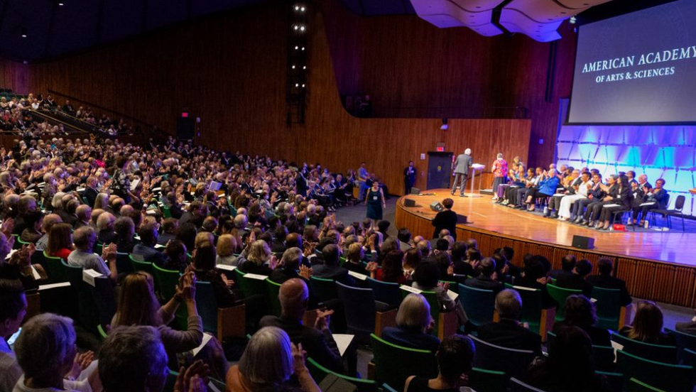 Audience watches a group of seated people on a stage with a projected screen reading "American Academy of Arts & Sciences"