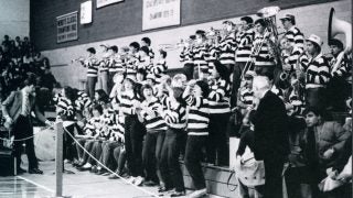 A black-and-white photo of the Pep Band, who are wearing striped long-sleeve shirts, standing on bleachers and cheering on a basketball game in 1984