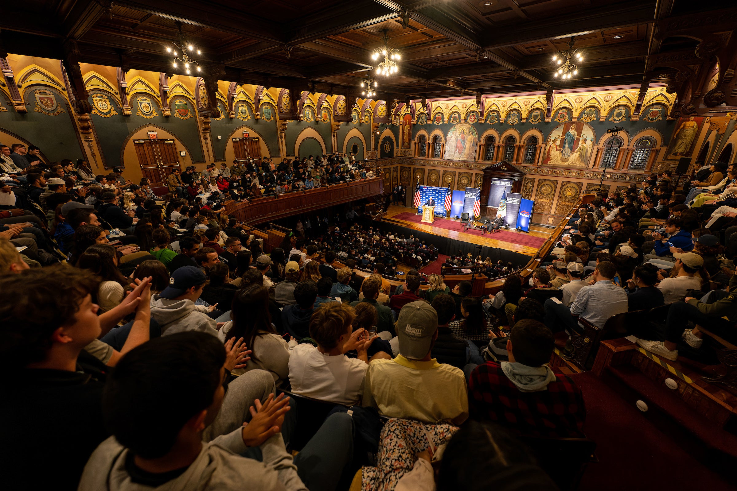 Large audience in an ornate auditorium looks at banners set up on a stage