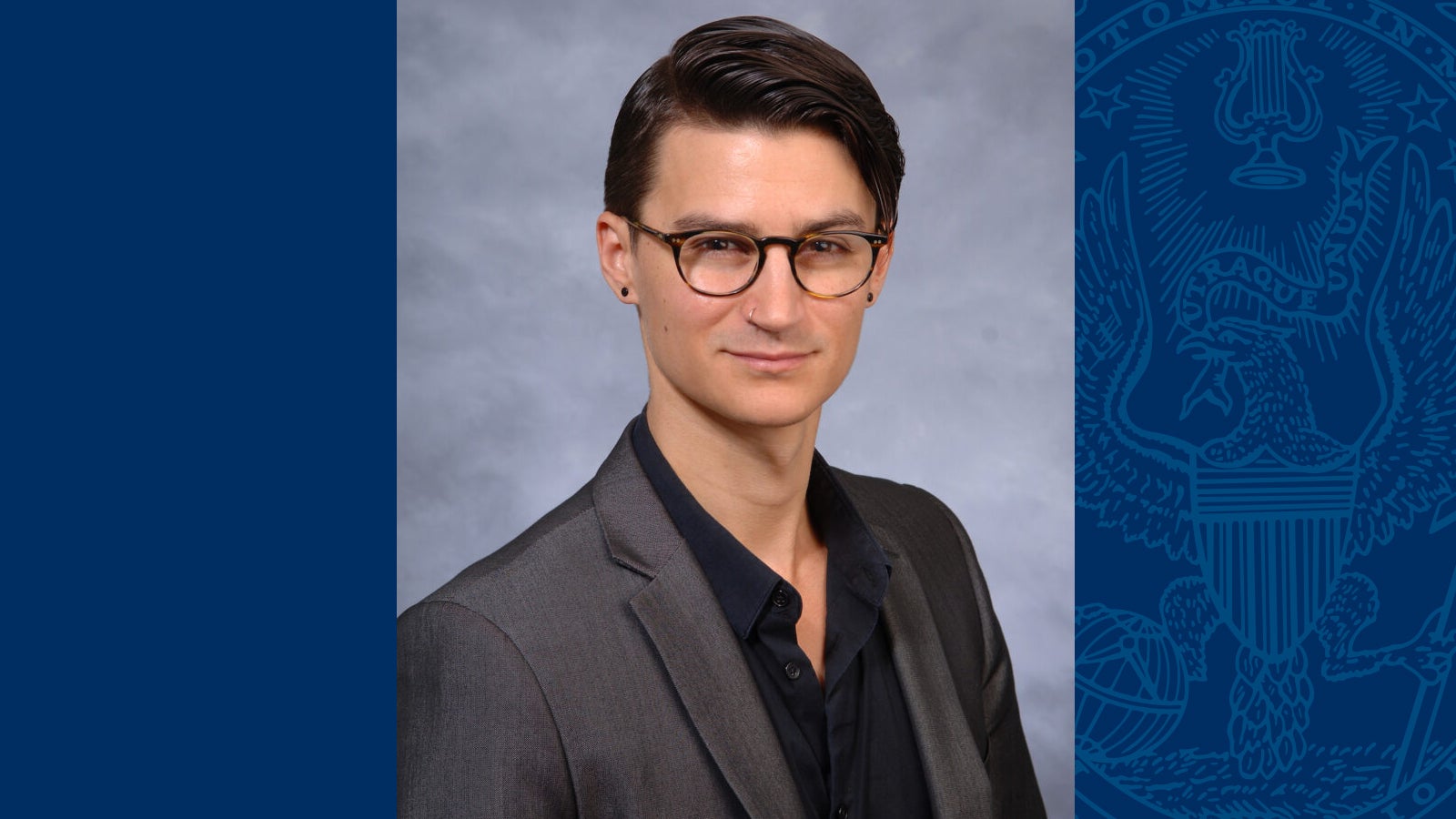 Joel Reynolds wears a gray blazer and black collared shirt, classes, and earring in a headshot against a gray background