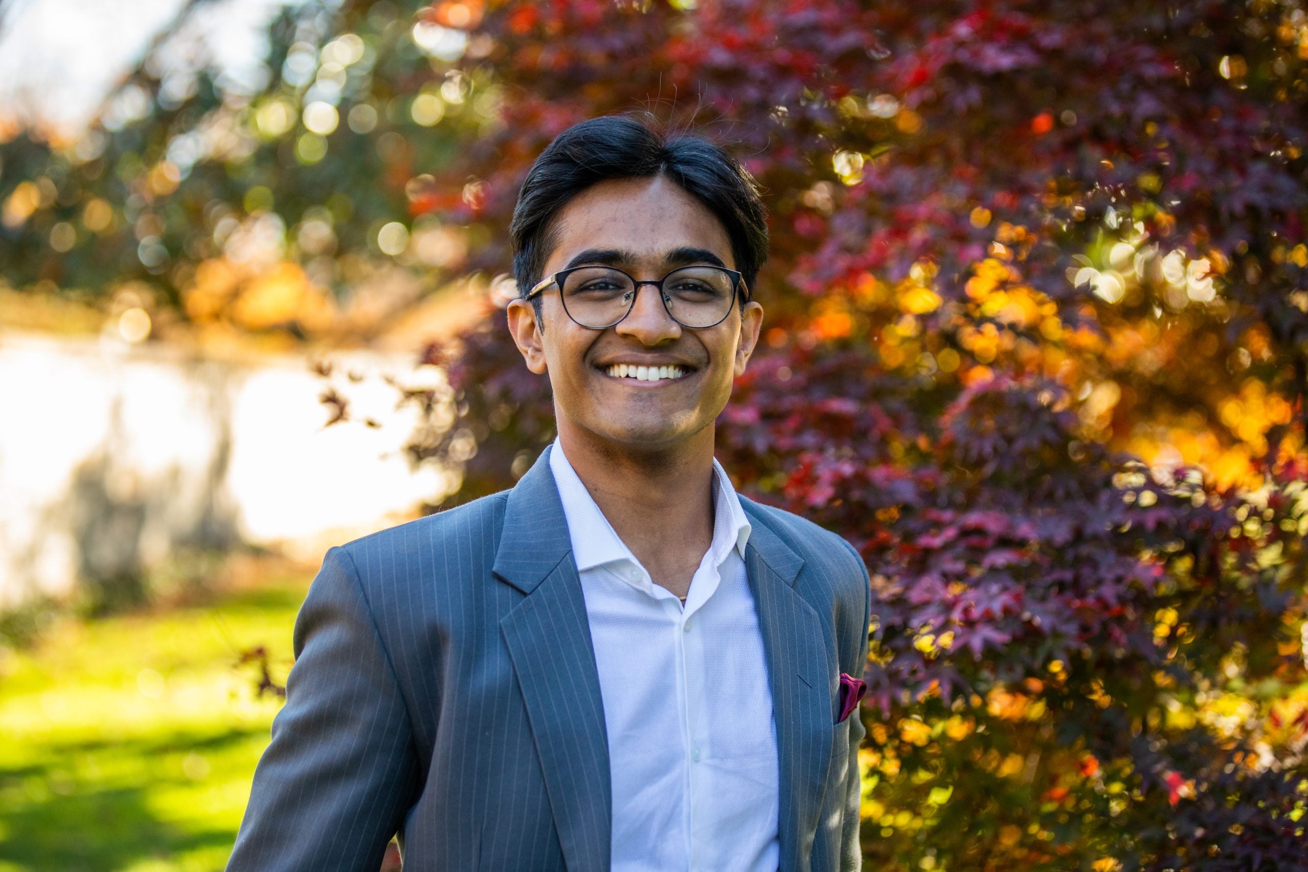 Atharv Gupta wears a white collared shirt and pinstripe gray blazer in front of a red, orange and yellow leaved tree