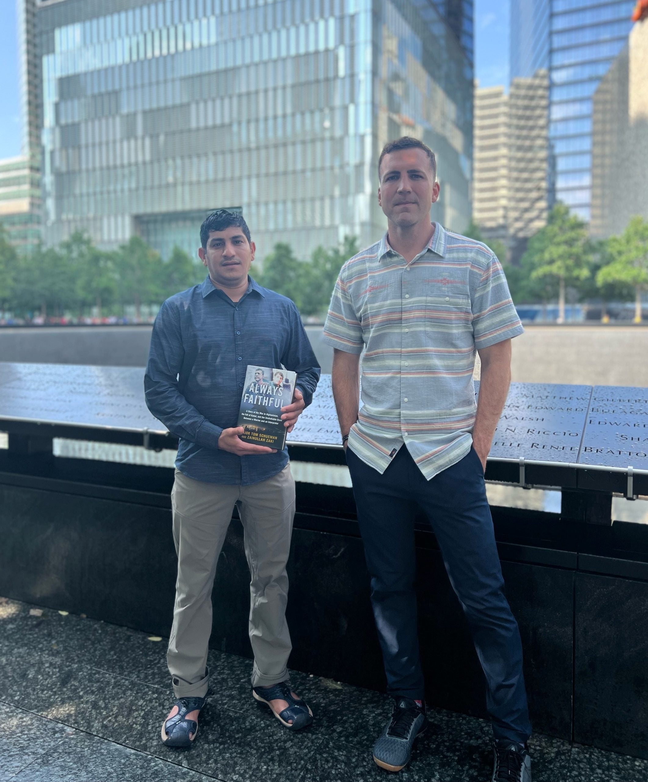 Two men stand next to each other in front of blue river and skyscrapers. The man on the left holds a book and is wearing a blue long-sleeved shirt and khakis. The man on the right wears a short-sleeve button-down and jeans.