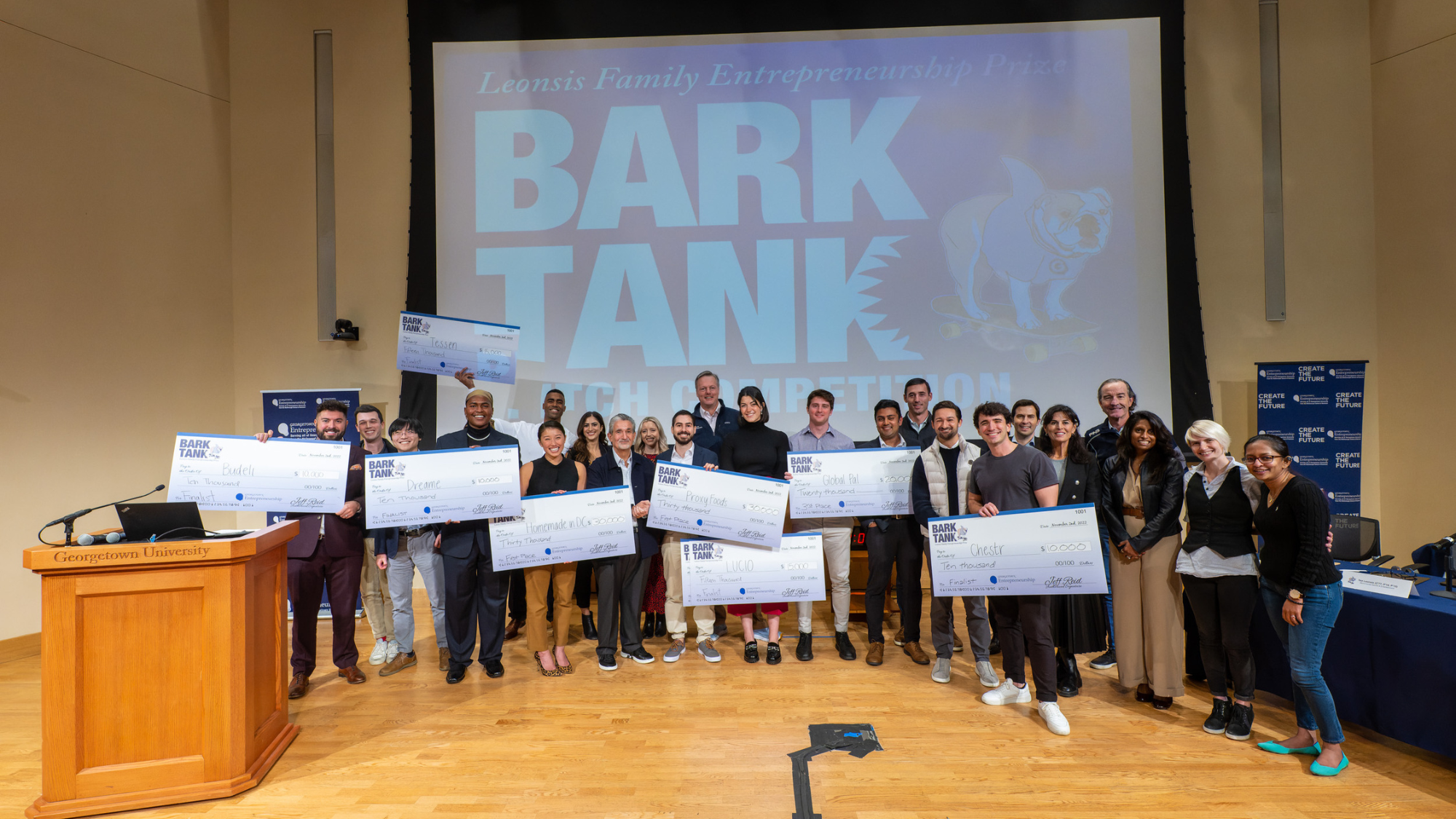 A group of students stand on stage holding giant checks with a screen behind them that says "Bark Tank"