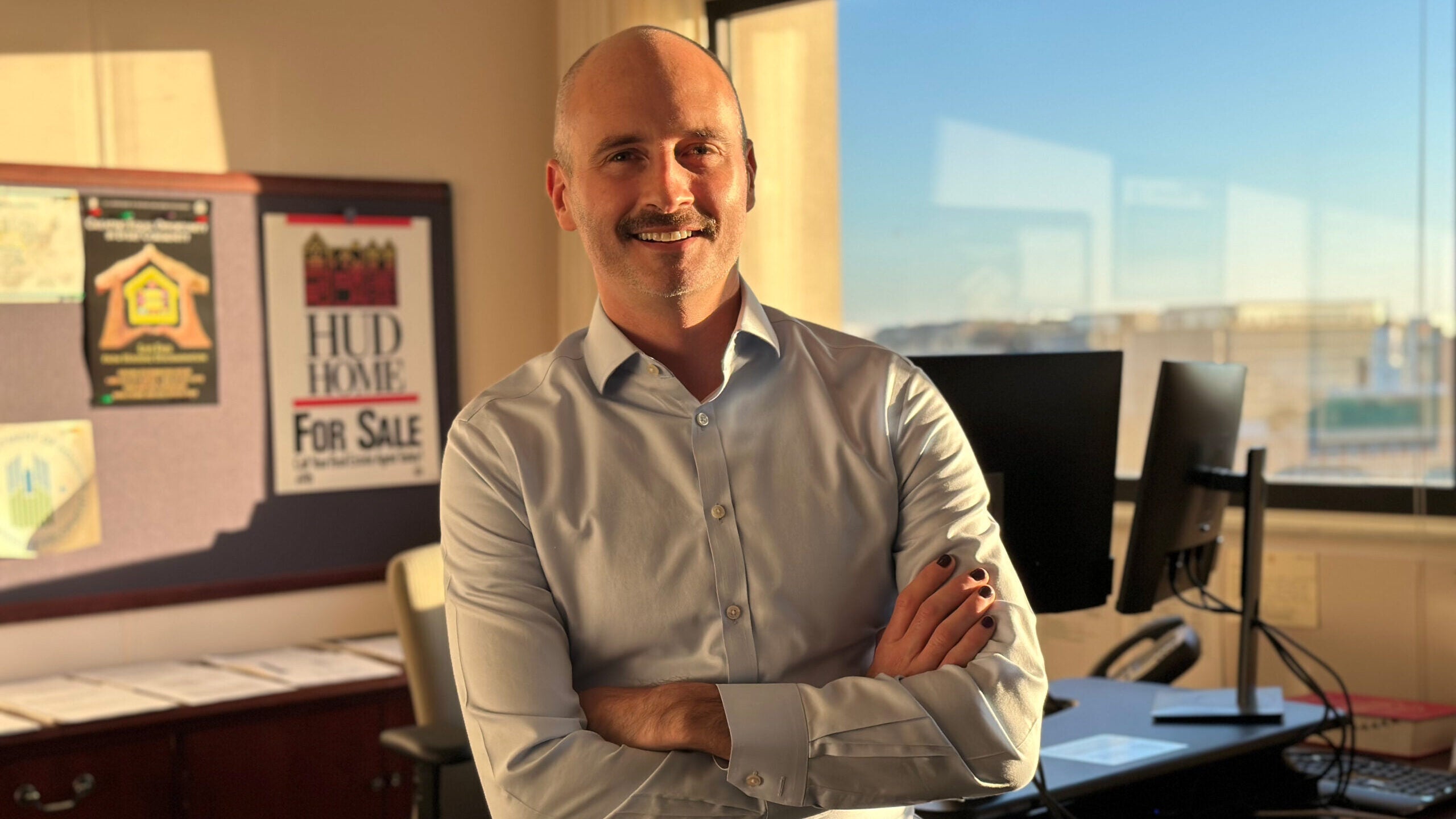 Brian McCabe (SFS'02) stands in his office wearing a blue button-down. Behind him to his right is a window with blue skies, and behind him to his left are posters that say "HUD" on them.