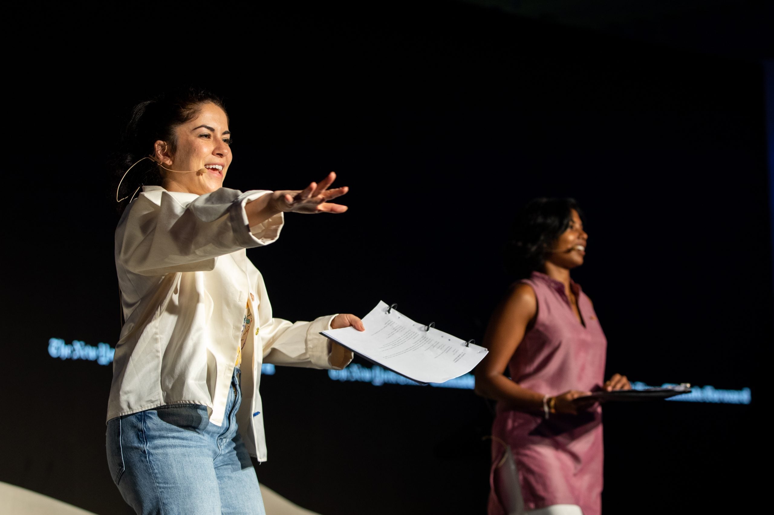 Two women stand on a stage with a black background at the New York Times' Climate Forward program. One woman wears a white jacket and blue jeans, and other other woman, on the right, wears a pink dress.