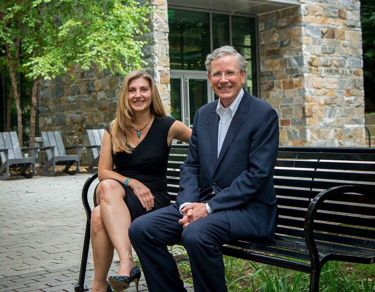 Sheila McMullan (left) wears a black dress and John Monahan (right) wears a suit as they sit on a bench in front of a stone building